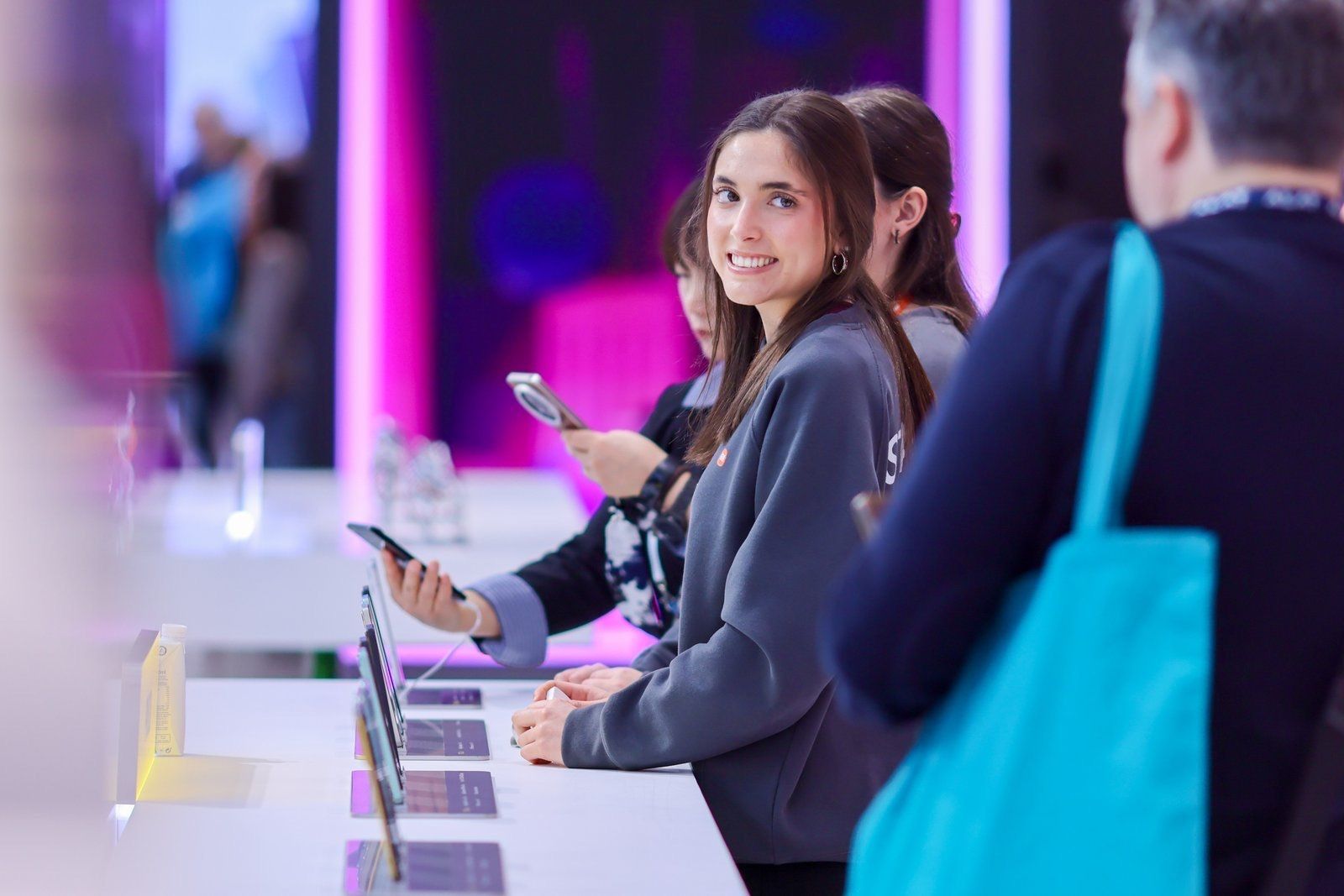 Azafata sonriente ante un expositor de teléfonos iluminado en el stand