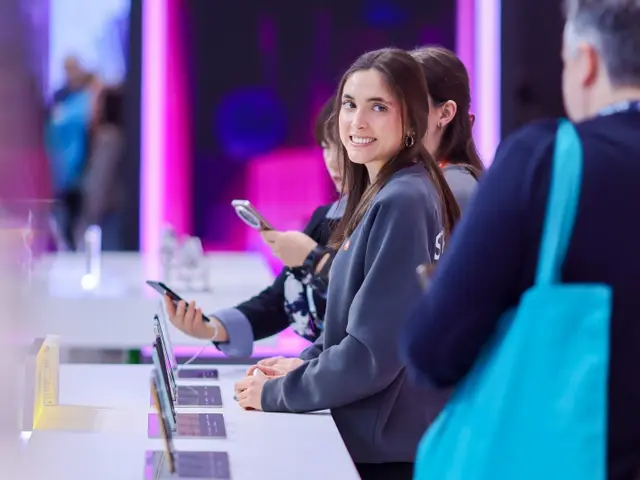 Azafata sonriente ante un expositor de teléfonos iluminado en el stand